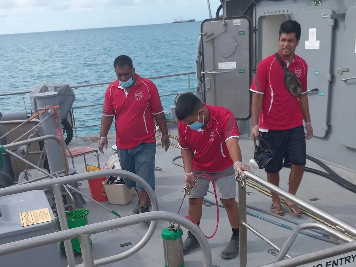 Three Terminix staff in red shirts on the open deck of a patrol boat, one crouching to spray from a hand-pumped canister, with the sea behind them
